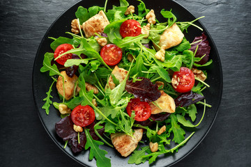 Fresh salad with chicken breast, arugula, nuts and tomatoes on black plate in a wooden table.
