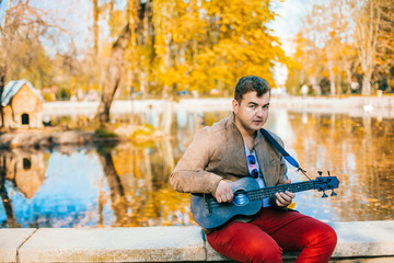 Young man are playing bass ukulele on the park autumn park near lake. Basking  guy in the street.