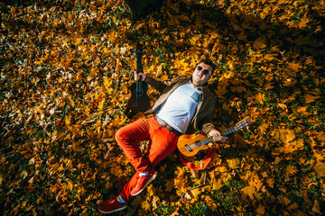 Young guy musician lying on the yellow leaf with two ukulele guitars in autumn sunny day. Man In red pants and pink glasses. 