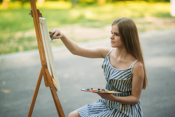 Beautiful girl draws a picture in the park using a palette with paints and a spatula. Easel and canvas with a picture.