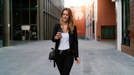 Photo of young woman reading messages on a smartphone while standing on urban background. Blonde caucasian female is surfing the web on a mobile phone while walking the business centre area.