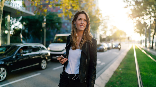 Photo of caucasian female listening to music on a smartphone while standing on street background. Model look blonde female is surfing the web on a mobile phone while walking the city center.