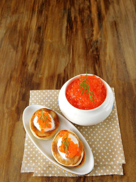 Red caviar in a white ceramic bowl and pancakes with caviar on a wooden table