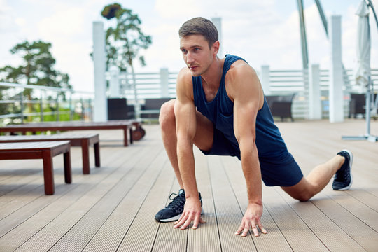 Portrait of muscular sportsman working out on wooden terrace outdoors stretching legs during warm up
