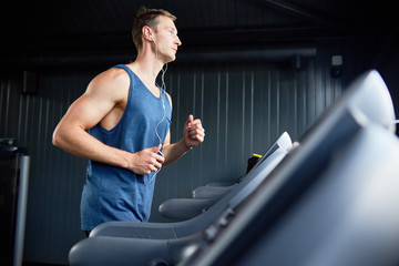 Side view portrait of muscular young man running on treadmill in modern gym during cardio workout,...