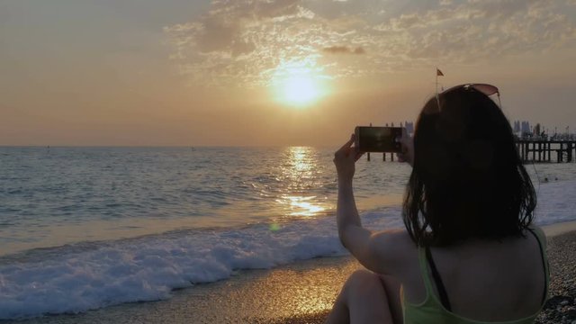 An attractive woman, a brunette is taking pictures on the phone with a beautiful view of the sea and a colorful sunset.