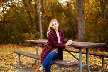 young blond woman sitting on park bench in autumn with coffee
