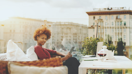 Charming young naughty black woman sitting on the wicker bengh outdoors in roof restaurant with digital tablet; biracial teenage female freelancer in street cafe with digital pad, laptop, and teapot