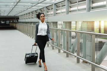 Fototapeta na wymiar Beautiful businesswoman walking inside public transportation station with suitcase, front view