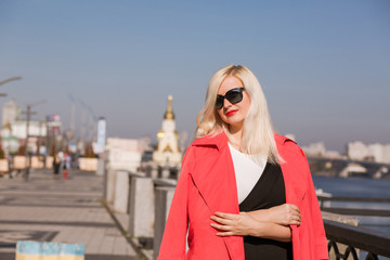 Obraz premium Stylish blonde model wears sunglasses, posing in red cloak on a blurred bridge background