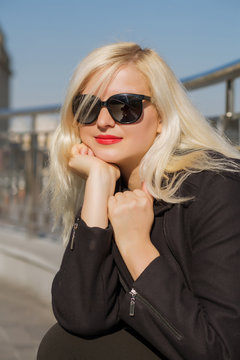 Closeup portrait of blonde model posing in black cloak, wears sunglasses  on a blurred city background