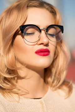 Fashion portrait of glamor blonde model wears clear glasses with bright makeup posing in sun light