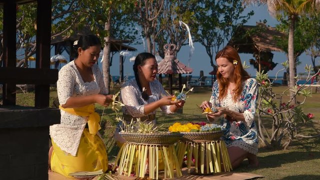 Two balinese women showing to young caucasian woman how to make canang sari offerings, sitting outdoors on green lawn. Shot with Sony a7s and Atomos Ninja Flame on sunny summer day in Bali, Indonesia.