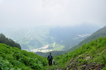 Obraz premium Two young men, come back from a campaign, climbing down a mountain among green vegetation and against the background of clouds and the town is below.
