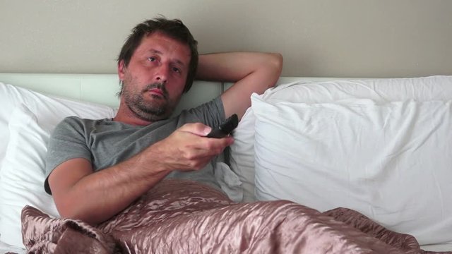 Man using TV remote controller in bedroom, laying alone in bed and watching television or movie series