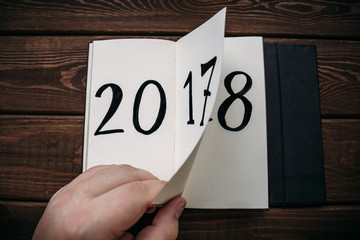 New Year 2018 is coming concept. Hand flips notepad sheet on wooden table. 2017 is turning, 2018 is opening