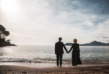 Fototapeta premium Rear view of happy couple standing against a beach side in summer. Two lovers in vacation in an idyllic nature scene sharing positive feelings and emotions. Magic moments of loving hearts.