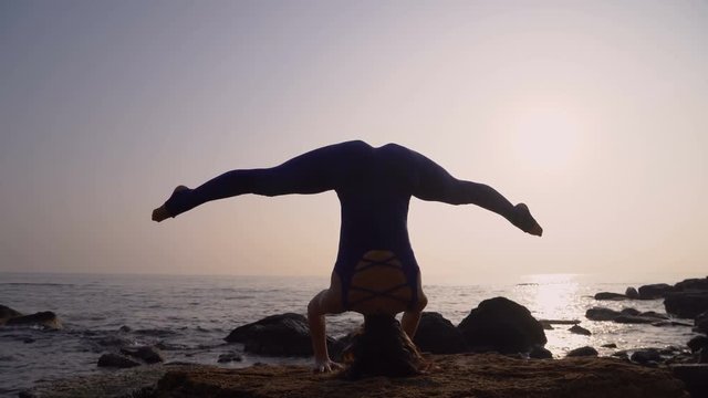 Young woman in bodysuit practicing yoga on the beach above sea at amazing sunrise. Fitness, sport, yoga and healthy lifestyle concept. Girl doing stand on head and twine