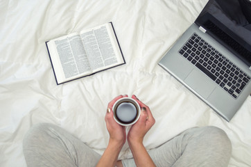 Mão de mulher segurando xícara de café, livro e notebook na cama, visto de cima.