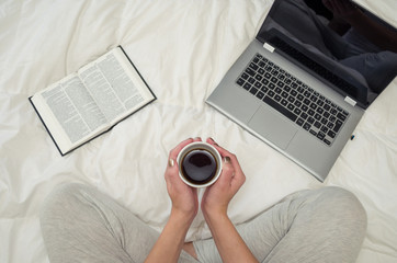 Mão de mulher segurando xícara de café, livro e notebook na cama, visto de cima.