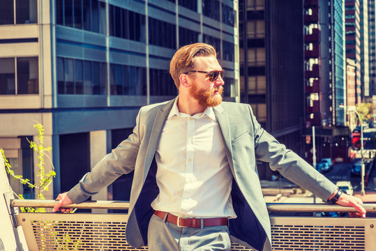 American Man with beard, mustache working in New York, wearing cadet blue suit, white shirt, sunglasses, standing by railing on balcony, facing street with high buildings under sun, waiting to meet.