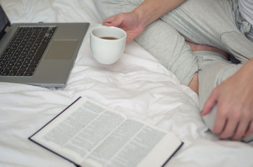 Mulher bebendo café na cama com livro e computador, vista lateral