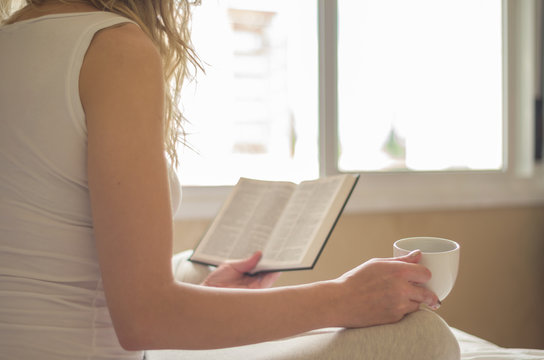 Jovem mulher caucasiana lora lendo livro, bíblia, bebendo café, chá na cama com raios de sol pela janela, manhã cedo.