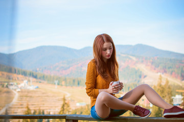 Cute and nice young woman drinking coffee at terrace on a sunny morning in mountain. Pretty lady in stylish casual yellow sweater and shorts. Warm soft cozy image, breakfast on the balcony  