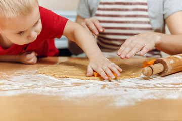 Little kid boy helps mother to cook Christmas ginger biscuit in light kitchen. Happy family mom 30-35 years and child 2-3 roll out dough and cut out cookies at home. Relationship and love concept