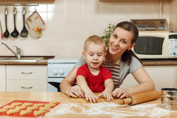 Obraz na płótnie Canvas Little kid boy helps mother to cook Christmas ginger biscuit in light kitchen. Happy family mom 30-35 years and child 2-3 roll out dough and cut out cookies at home. Relationship and love concept