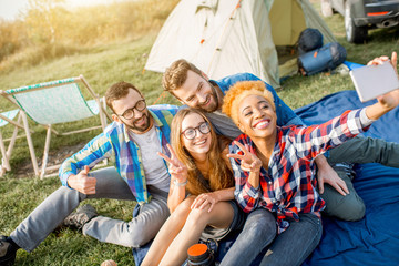 Multi ethnic group of friends dressed casually having fun making a selfie photo together during the outdoor recreation with tent, car and hiking equipment near the lake