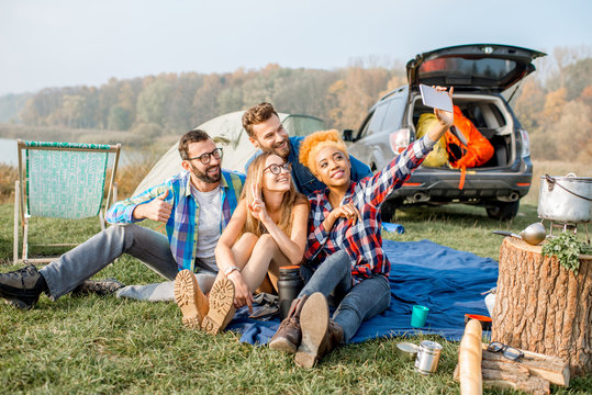 Multi ethnic group of friends dressed casually having fun making a selfie photo together during the outdoor recreation with tent, car and hiking equipment near the lake