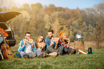 Multi ethnic group of friends having a picnic, eating watermelon, sitting in a row at the camping with tent, car and hiking equipment near the lake