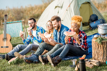 Multi ethnic group of friends having a picnic, eating watermelon during the outdoor recreation with tent, car and hiking equipment near the lake