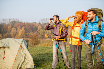Multi ethnic friends in colorful jackets hiking with backpacks, standing near the camping on the green lawn
