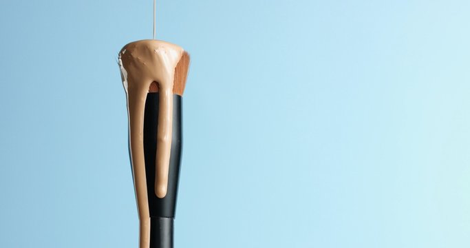 Pouring bb cream or foundation on the bristles of a professional make up brush with black handle isolated on blue