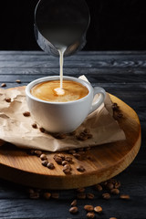 Pouring milk in coffee. Cup with cappuccino on wooden plate