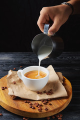 Pouring milk in coffee. Cup with cappuccino on wooden plate
