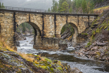 Fototapeta na wymiar A beautiful historic stone bridge crossing the river in central Norway. Colorful autumn landscape.