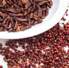 Dried small red peppers in close-up on a white plate and pink pepper grains on a white table.