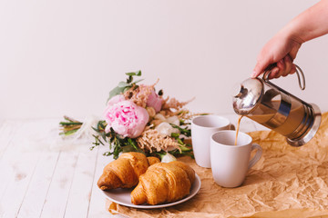Delicious healthy breakfast with two fresh flaky croissants served on a plate with two cups of coffee and a bouquet of summer wildflowers on kitchen table. Woman's hand pouring coffee