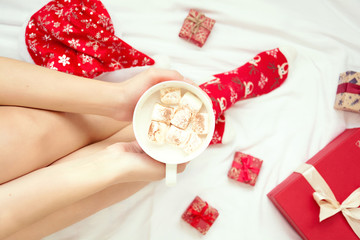 Woman taking mug with hot chocolate and marshmallow on white bed sheet. Overhead shot.
