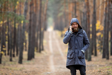 A man in a gray jacket stands on the road in the middle of a pine forest and talks on the phone 