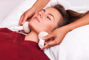 Herbal ball face massage in ayurveda spa. Female massagist with young woman in wellness center.