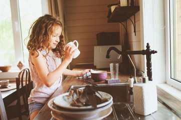 child girl helps mother at home and wash dishes in kitchen. Casual lifestyle in real interior. Teaching kids to do house work.