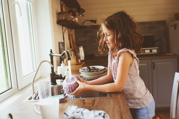 child girl helps mother at home and wash dishes in kitchen. Casual lifestyle in real interior....