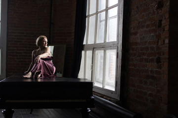 A woman in a pink silk dress sits on a piano and looks out the window