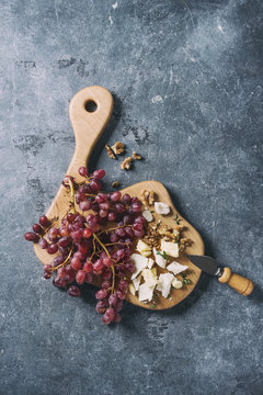 Wooden serving board with fresh red grapes, walnuts, goat and cheddar cheese, with cheese knife over blue texture background. Top view, copy space. Toned image