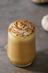 Glass of spicy pumpkin latte with whipped cream and cinnamon standing on gray kitchen table with decorative white pumpkins. Coffee beans and spices above. Atmospheric breakfast