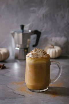Glass of spicy pumpkin latte with whipped cream and cinnamon standing on gray kitchen table with coffeepot and decorative white pumpkins. Coffee beans and spices above. Atmospheric breakfast